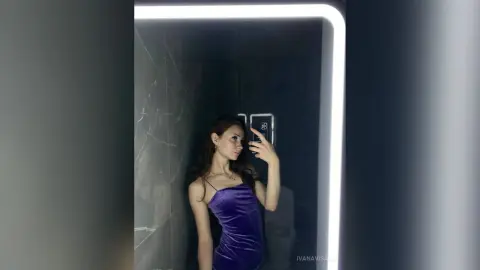 Young woman in a purple velvet dress taking a selfie in a modern bathroom with bright LED lighting.