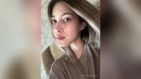 Young woman with long blonde hair wearing a brown ribbed sweater in a soft, natural indoor setting.