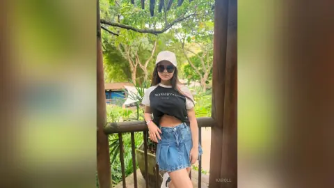 Young woman in a black graphic t-shirt and denim skirt posing outdoors in a lush green garden setting.