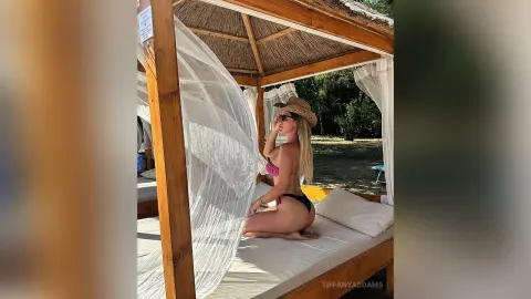 TiffanyAddams poses in a black bikini and straw hat while kneeling on a white bed inside a wooden cabana with white netting.