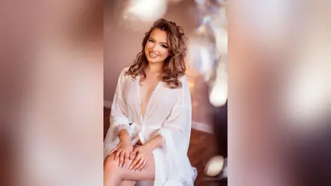 Smiling young woman in a white lace robe posing in a warm, glowing indoor setting.