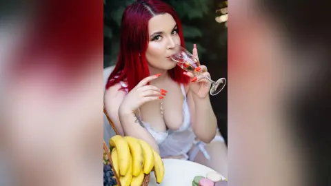 Red-haired woman in white lace lingerie sipping wine at a romantic outdoor brunch with fruit.