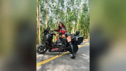 Stylish woman in black leather outfit posing on a black scooter on a scenic forest road.