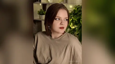 Young woman with dark hair and cross earrings wearing a beige t-shirt in a cozy indoor setting.