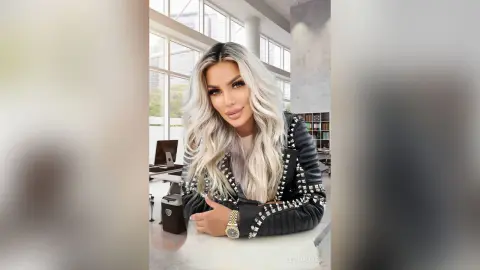 Smiling blonde woman in a black studded leather jacket sitting in a bright modern office setting.