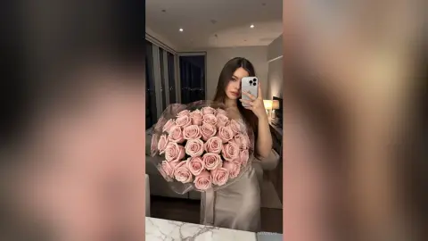 Young woman in a silver satin dress holding a large bouquet of pink roses for a romantic indoor selfie.
