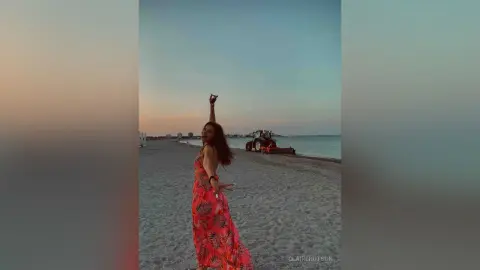 Joyful woman in a red floral maxi dress raising her arms on a peaceful beach at sunset.