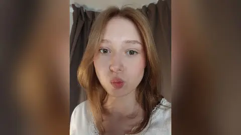 EvelinaMoorina pouts her lips at the camera while wearing a white lace top in front of brown curtains.