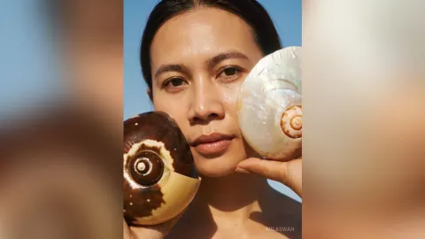 MilaSwan — MilaSwan holds large seashells against her face under a clear blue sky.