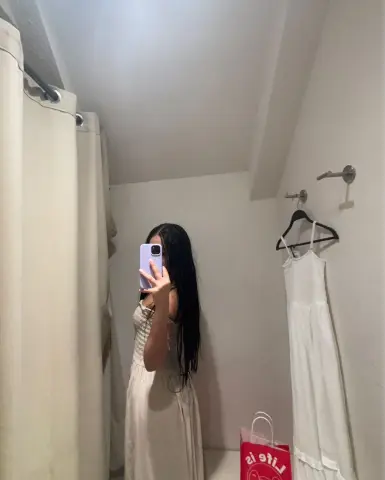 Young woman in a white maxi dress taking a mirror selfie in a minimalist cream-colored room.
