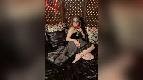Mysterious woman in a black lace dress and silver mask holding a red rose on a dark satin bed.