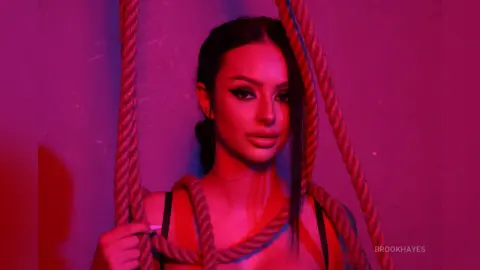 Beautiful woman with dark hair posing amidst thick ropes under moody pink and purple neon lighting.