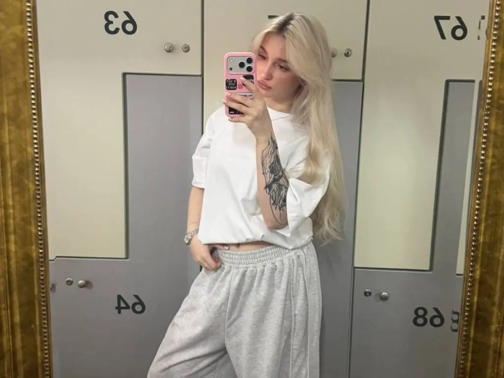Blonde woman in white t-shirt and grey sweatpants taking a mirror selfie in a fitting room.