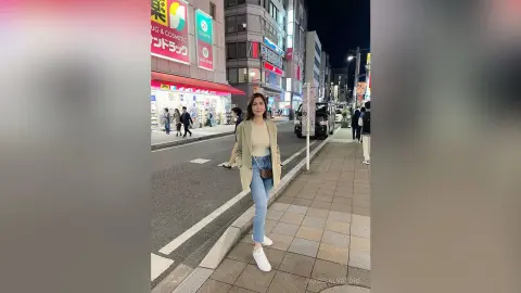 Stylish woman in a beige blazer and blue jeans walking on a city street at night near glowing storefronts.