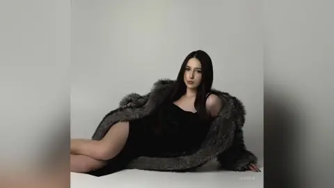 Elegant woman in a black velvet dress and oversized faux fur coat posing against a minimalist white background.
