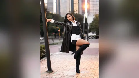Stylish woman in a black leather trench coat and boots posing confidently on a city street at sunset.