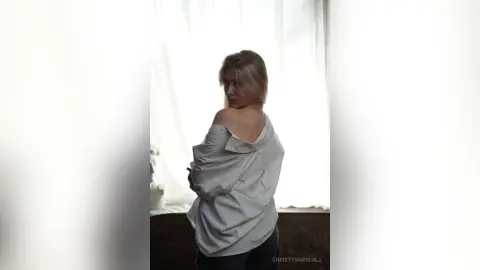 Blonde woman in an oversized white button-down shirt posing by a bright window in a soft, airy setting.