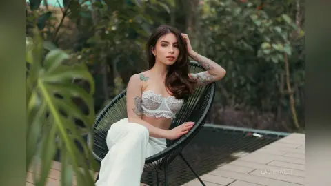 Tattooed woman in white lace lingerie and linen pants relaxing on a patio chair in a lush garden setting.