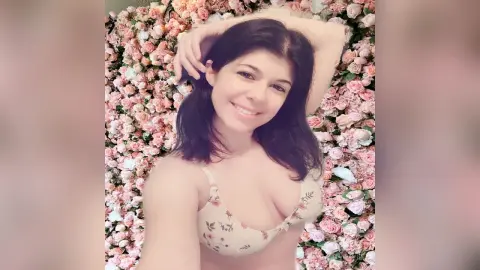 Smiling woman in a floral print bikini top posing against a romantic pink rose flower wall.