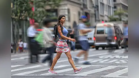 Stylish woman in a blue crop top and patterned mini skirt walking across a busy city street crosswalk.
