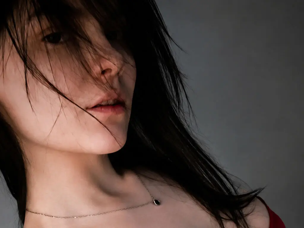 Close-up portrait of a woman with dark hair and a silver necklace against a moody gray background.