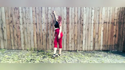 Young woman in a black crop top and red trousers posing against a rustic wooden fence in natural light.