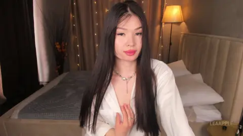 Young woman with long dark hair wearing a white shirt sitting on a bed in a cozy, dimly lit bedroom.