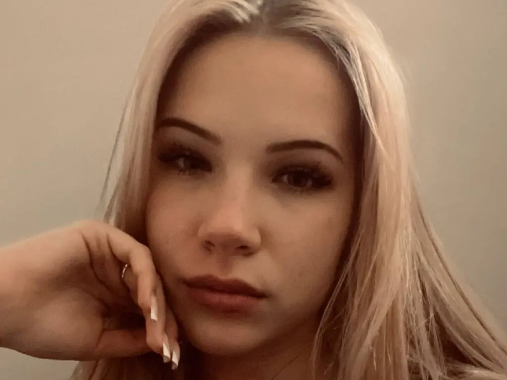 Close-up portrait of a blonde woman with natural makeup posing with a thoughtful expression against a neutral background.