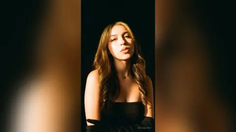 Young woman in a black strapless top posing under warm cinematic lighting against a dark background.