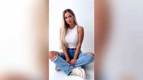Blonde woman in a white tank top and ripped blue jeans sitting cross-legged against a white background.
