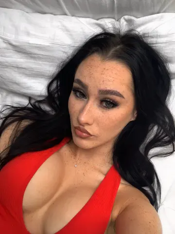 Young woman with freckles and long dark hair wearing a red ribbed tank top relaxing on white bedding.