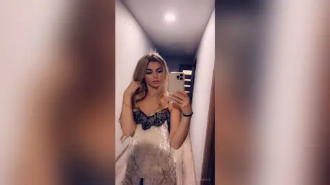 Glamorous blonde woman in a black lace cocktail dress taking a mirror selfie in a modern hallway.