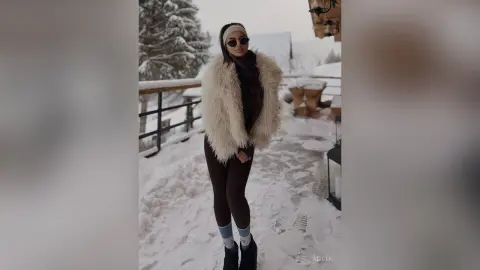 Stylish woman in a white faux fur coat and sunglasses posing on a snowy balcony in a winter landscape.