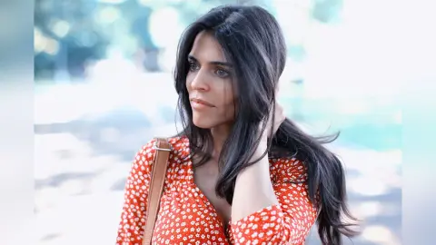 Thoughtful woman in a red floral blouse standing outdoors in soft natural light.