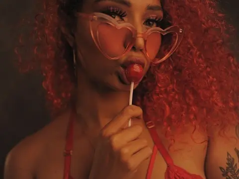 Glamorous woman with curly red hair wearing heart-shaped sunglasses and red bikini enjoying a lollipop.