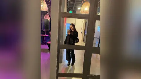Stylish woman in a black blazer taking a mirror selfie in a modern, dimly lit lounge with warm pendant lighting.