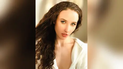 Beautiful woman with long wavy brown hair wearing a white top in a bright, soft-lit indoor setting.