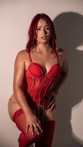 Red-haired woman in a red lace corset and stockings posing against a neutral wall in moody lighting.