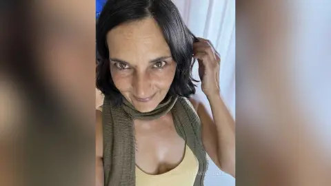 MichaelaBelle smiles while touching her dark hair, wearing a light yellow tank top and an olive green scarf.