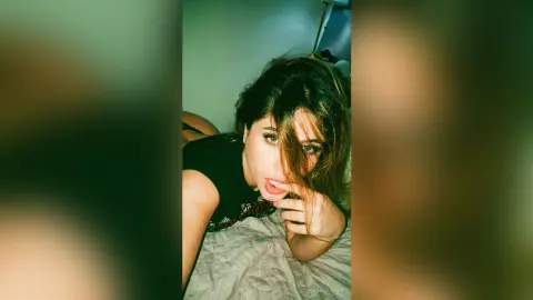 IrinaMolkova looks at the camera with her hair falling over her face while wearing a black t-shirt and lying on a bed.