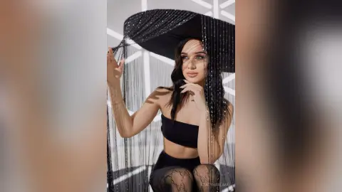 Elegant woman in a black strapless top posing under a shimmering black fringe curtain in a bright studio.