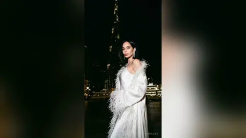 Elegant woman in a white feathered gown posing against the night skyline of Dubai near the Burj Khalifa.