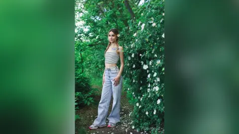 Young woman in a striped crop top and light blue jeans standing in a lush green garden with white flowers.
