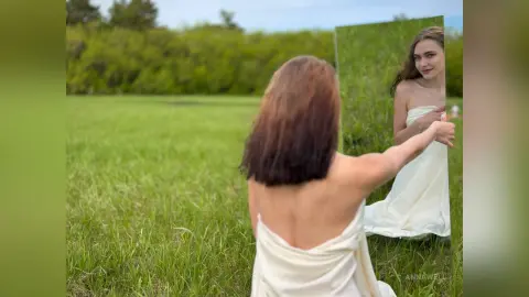 Woman in a white flowing dress looking into a mirror in a lush green meadow during a dreamy outdoor photoshoot.