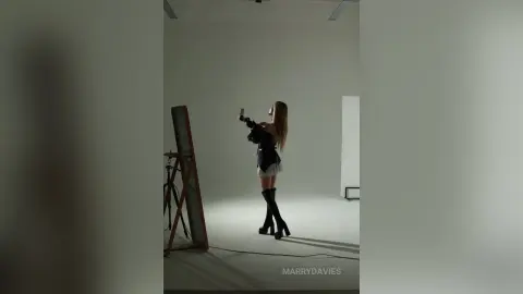 MarryDavies stands in profile against a white studio wall, wearing a black top and light skirt with black thigh-high boots.