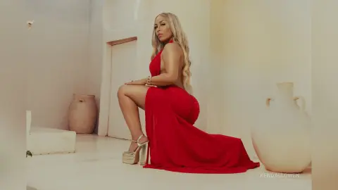 Elegant blonde woman in a flowing red evening gown sitting in a minimalist white Mediterranean room.