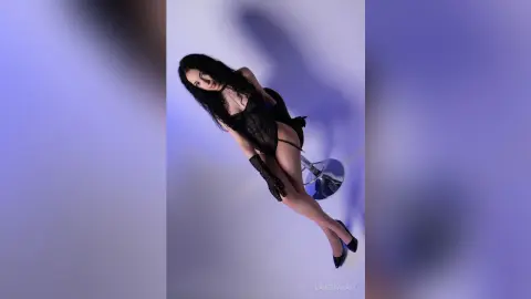 Elegant woman in a black lace bodysuit and gloves posing against a soft purple studio background.