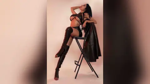 NicoleBella poses sitting on a black stool wearing a black leather bra and high-waisted bottoms with matching thigh-high boots.