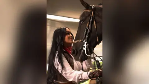 Beautiful woman in western equestrian outfit bonding with a dark brown horse in a rustic stable setting.