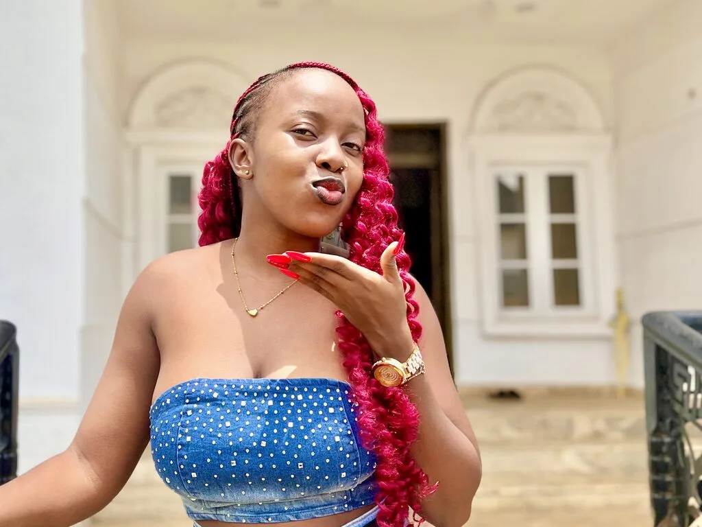 CynthiaOnyi — CynthiaOnyi pouts her lips while wearing a blue polka-dot tube top and a gold watch against a white building background.